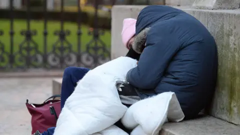 Two homeless people sleeping on the plinth of a statue. They are dressed in thick coats and are holding a large duvet to keep warm.
