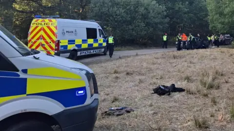 Norfolk Constabulary Two police vans, one parked on grass and the other on the road. Multiple police officers in high-visibility jackets are gathered around two cars and a couple of non uniformed people. There are some discarded clothes on the grass. 