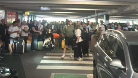 Dozens of people stand in a car park by a zebra crossing with their luggage in among parked cars.