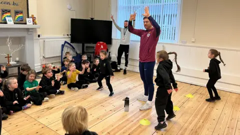 A man in sports gear and a burgundy England football top leads a group of schoolchildren in an exercise routine. Some of the children are standing up and jumping whereas the majority are sitting down and watching