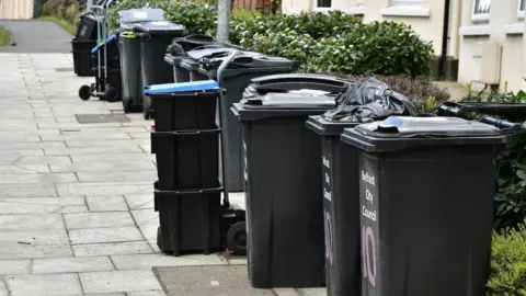 Pacemaker A row of black bins. Some of them are open with black bin bags overflowing. There is a stack of smaller black bin boxes. On the front of the bins it says Belfast City Council in white writing.