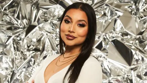 A woman with long dark hair and wearing a white stop, turned to the side and looking straight at the camera against a silver backdrop.