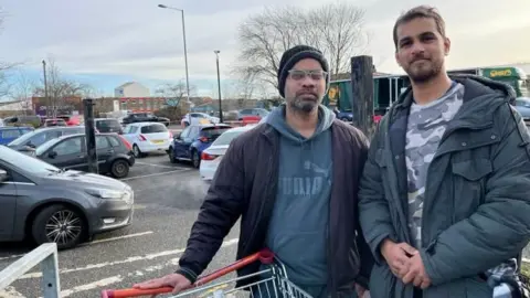 Two men in dark jackets stand next to a trolley in a car park. One has his hand on a trolley. They are both wearing coats. 