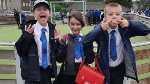 Ryan Quinn Aaron and Ryan in their school uniforms in the school playground pull silly faces for the camera. A female friend smiles in between the two of them. Their school ties are a light blue with a dark blue check pattern. The girl carries a bright red satchel.