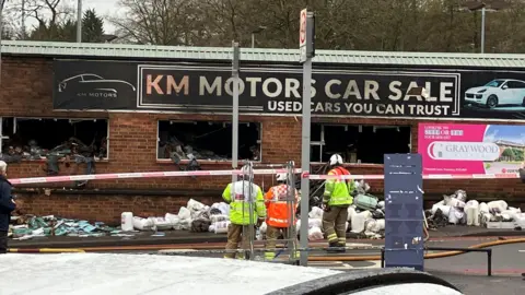 Three firefighters outside a building which has a red and white cordon in place. The building has windows which are burnt and damaged and there are piles of rolled materials outside. The sign about the building says 'KM Motors Car Sale Used cars you can trust'. 