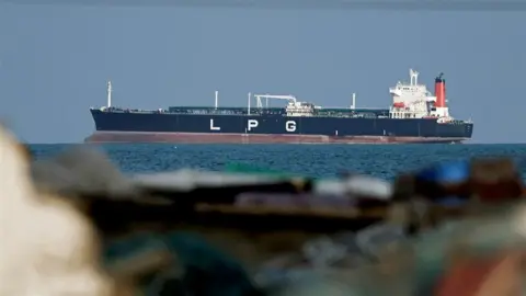 Close up of a ship on blue water in the background. In the foreground are rocks
