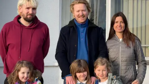 Three parents - one man with blond hair and a dark beard, another man with red hair and a beard and a woman with long dark hair stand in front of three girls with long hair