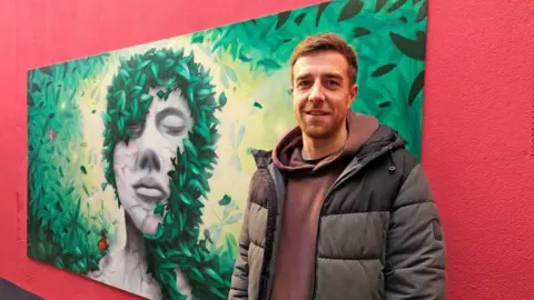 Newcastle City Council Artist Mark One87 standing by his work. He has short brown hair and stubble. he is standing a painting of a grey woman covered in leaves, which are growing in from the sides of the painting.