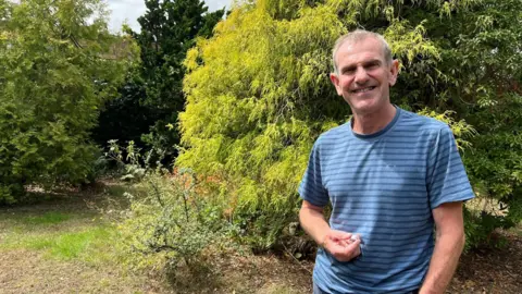 Gregory Quinn stands in his front garden with a thick green hedge behind him. He is wearing a blue striped t shirt and is smiling at the camera. 