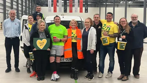 Group of people stand in front of a van and smile at the camera. One man - Mike - wears a green shirt and holds a red cup, he's stood next to a woman with a high-vis jacket. People hold collection buckets and a sign which says 'thank you'