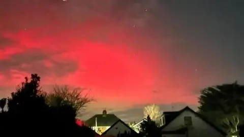 An amateur photograph of the Northern Lights. There is a pink light in the sky above two houses.