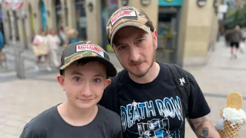 Andy Naylor on the right and son Ronan on the left. They are wearing matching camo caps with a red white and black logo. Andy is holding an ice cream and is wearing a black t-shirt with blue writing on it. He also has a black bag pack on. Ronan is wearing a black t-shirt. The background behind is blurry.
