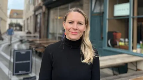 BBC Ella dressed in a black top standing in a narrow pedestrian street lined her café. Outdoor tables and signage are visible, and the sky appears partly cloudy.

