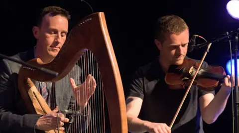 Two men sitting side by side as they play their instruments. Mr Rooney on the left plays his brown wooden harp, he has short dark hair and is wearing a dark grey suit with a patterned tie. On the right is Mr Byrne with short brown hair and facial hair who is wearing a black t-shirt and is playing a fiddle. Behind then both is a black backdrop and two coloured stage lights.