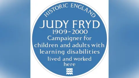 Historic England Blue plaque with the words "Judy Fryd, 1909-200. Campaigner for children and adults with learning disabilities lived and worked here", in white lettering.