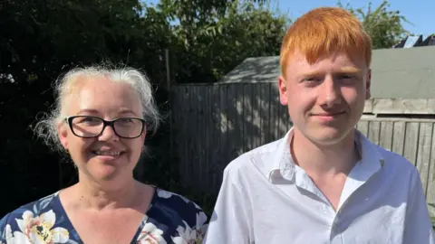 Kitty and George standing side by side outdoors in a sunny backyard. Kitty is wearing a dark blue dress with a light-colored floral pattern and glasses, while George is wearing a white short-sleeved button-up shirt. Behind them is a wooden fence, a small shed and trees.