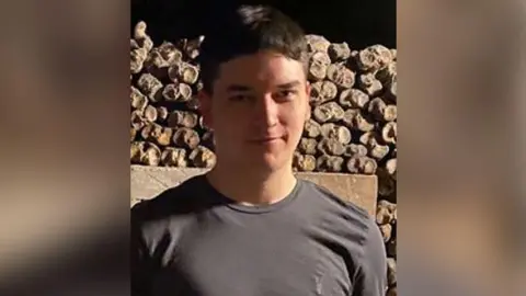 Tom Dingle with short brown hair wearing a grey t-shirt stands in front of a stone wall in the open air. It is evening. He is smiling.