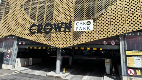 The outside of a multi-storey car park, a sign reads Crown Car Park. A yellow facade is on the outside walls of the building. Two barriers block two roads into the car park. 