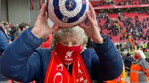 Peggy Pickavant is holding the match ball following a game at Anfield. She has raised the ball above her head and is standing in the Kop. She is wearing a Liverpool FC mask across her face and has a scarf around her neck.
Credit: Peggy Pickavant