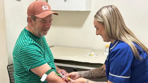 Morris Weightman in a cap and a green t-shirt having a needle put into his arm as a nurse in blue uniform takes his blood. Both are smiling.
