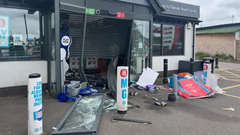 A Co-op shopfront has been smashed. There is a door with broken glass on the floor, a half opened black shutter on the front of the store, debris, litter and rubbish is seen strewn along the floor