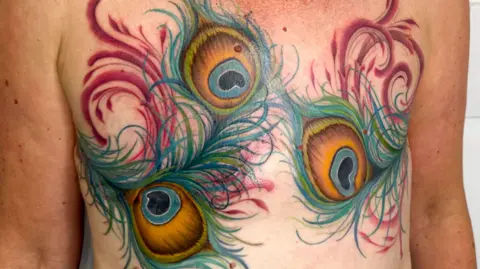 Tanya Buxton A masectomy tattoo - covering someone's chest - of three large peacock feathers with swirl details. A faint outline of masectomy scars can be seen beneath.
