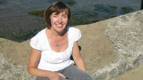 A woman sits on a concrete ledge beside clear, gently moving seawater, smiling toward the camera on a sunny day.