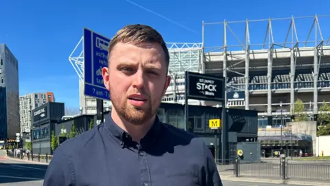 Daniel Mulligan. He has short brown hair and a beard and is wearing a blue shirt. He is standing in front of St James' Park.