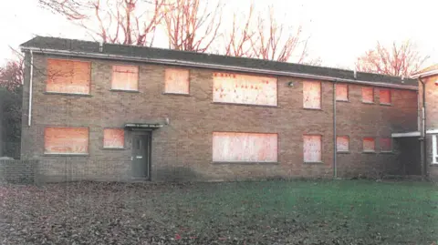 West Yorkshire Police A boarded up two-storey building with grass covered in leaves in the foreground.