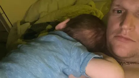 A young boy with dark hair and in a blue shirt lies asleep on the naked chest of a man who is looking at the camera.