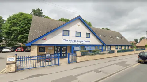 Google The exterior of a modern GP surgery with a large triangular shaped entrance and blue trim.