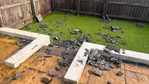 Roof slates are smashed in a garden. Debris stretches from the patio and all over the grass. 