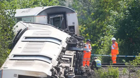 Three derailed train carriages lie on their sides on a railway embankment near Lewes in East Sussex. Rail workers in orange high-visibility clothing and a police officer are beside the track as investigators examine the scene. One carriage doorway is visible, showing damage where the train separated during the derailment.