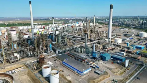 A drone view of an oil refinery, which includes a cluster of metal structures and storage containers on a concrete and asphalt base. A number of tall slim chimneys rising to the blue sky. Green fields and a river estuary can be seen in the background under a blue sky.