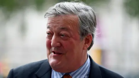Hannah McKay/Reuters Sir Stephen Fry, with grey hair and a clean shaven face, dressed in a blue and white striped shirt and tie and dark jacket. The backdrop is in soft focus, with some yellow, green and pink shades visible.