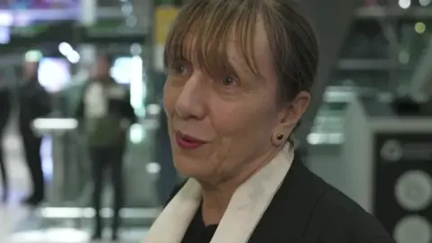 Gina is speaking to the camera. She has dark hair and a fringe. She is wearing a black jacket and a white scarf. 