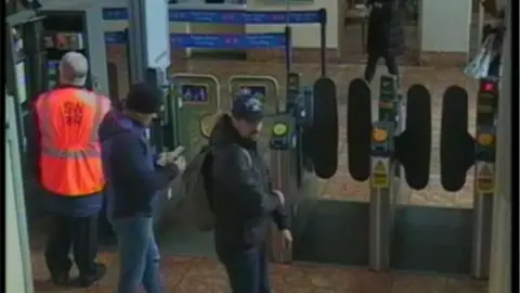 A CCTV image showing two men in dark jackets and hats next to ticket barriers at Salisbury Rail Station