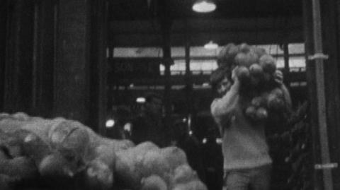 Black and white image of John Noakes humping some market goods over his shoulder walking towards a pile of the same product in front of him.