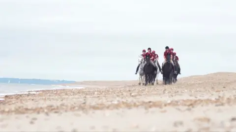 A group of black and white horses are gathered together running along the beach, their riders wear burgundy t shirts and black helmets