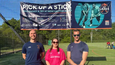 Three hockey coaches stand in front of the pitch. Finn and Harry stand either side of Emily, dressed in navy blue sports attire while Emily is wearing a pink hockey top.