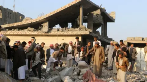 Yemenis inspect rubble. Men wearing mostly brown stand among the concretes ruins