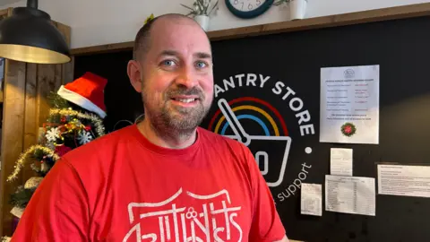 Richard Nunn is standing in front of a blackboard advertising the Salvation Army pantry store. He is wearing a red T-shirt with Salvation Army branding.