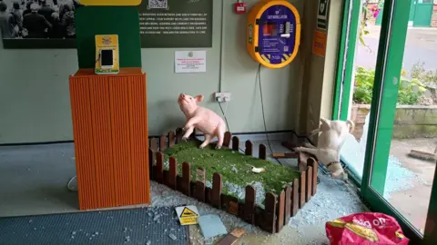 The image shows the scene of a break-in. It is in the foyer of a farm where a large glass door has been smashed and glass is all over the floor. A fake farm scene with a plastic pig and a goat is all thrown around and there is glass all over the artificial grass. 
