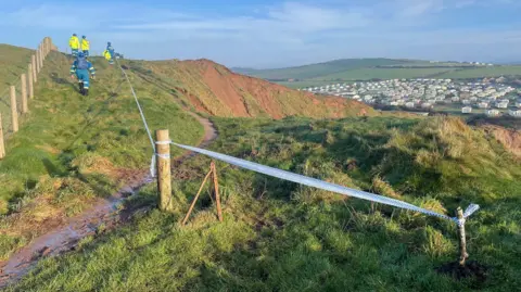 Todd Moore The top of the grassy cliff. Blue-and-white tape has been tied to posts, cordoning off the cliff edge. Several people in blue helmets and high-vis jackets are walking along the cliff-top.