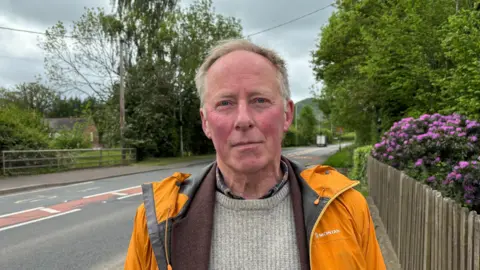 A man looks at the camera while wearing a golden orange raincoat and a brown jumper. There is a road behind him, a well as a purple flowered bush and trees