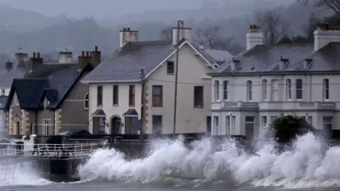 Waves break against a sea wall. Behind the wall and road and on the other side of the road houses that are grey, white and brown.