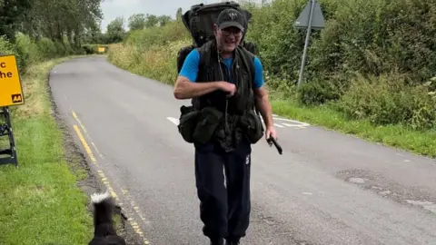 Philip Burlingham Philip wearing his old army gear and a blu t-shirt and dark cap walking on a road with his dogs