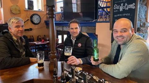 Carl Johnson is on the left of the photo. Mike Richards is in the middle of the photo and Marvin Odillard is on the right of the photo. They are sitting on stools at a bar. Carl is wearing a black jacket which is unzipped and has a checked shirt underneath. He has short grey hair. Mike has a black gilet jacket on with a white t-shirt underneath. He has short brown hair. Marvin is wearing a light green shirt which is unbuttoned with a black t-shirt underneath. He is bald but has dark stubble on his face.