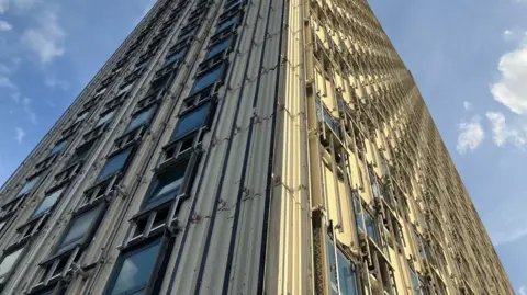 A close-up shot of Walbrook House, taken from below, showing the floors rising into the sky and rows of windows. It appears some of the concrete panels have been removed from the building's facade