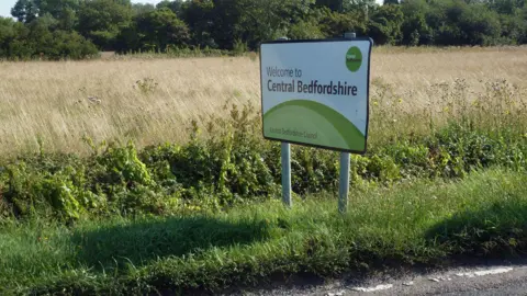 A sign reading: "Welcome to Central Bedfordshire", by the side of a road, with grass behind it in a field, a bush and more green grass. 